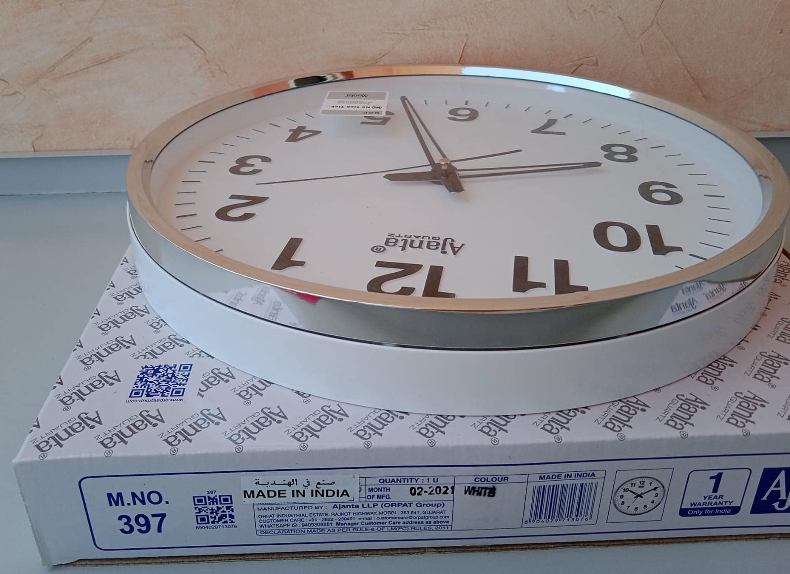 ساعة الحائط من أجانتا كوارتز. تتميز الساعة بمينا أبيض وإطار فضي أنيق يجمع بين التصميم العصري والأداء الموثوق. تعمل بتقنية الكوارتز لضمان دقة عالية في الوقت، كما أن الأرقام الواضحة تسهّل قراءتها من أي مسافة.